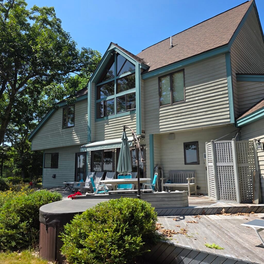 Close up view from porch of oceanside sprawling house with large bay windows with teal trim and stained gray shingles