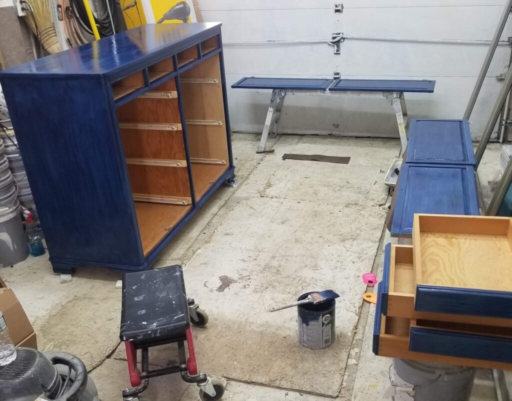 Cabinets, cabinet drawers, and cabinet doors are spread out throughout a workshop, freshly painted blue and set to dry before being reassembled