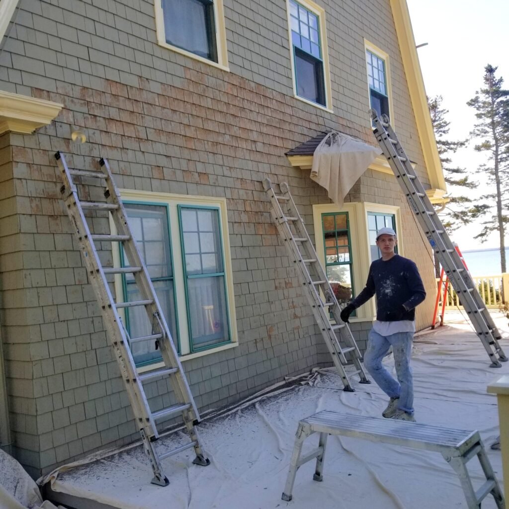 Crew member next to aluminum ladders by a large house in the process of scraping the siding tiles to remove peeling paint