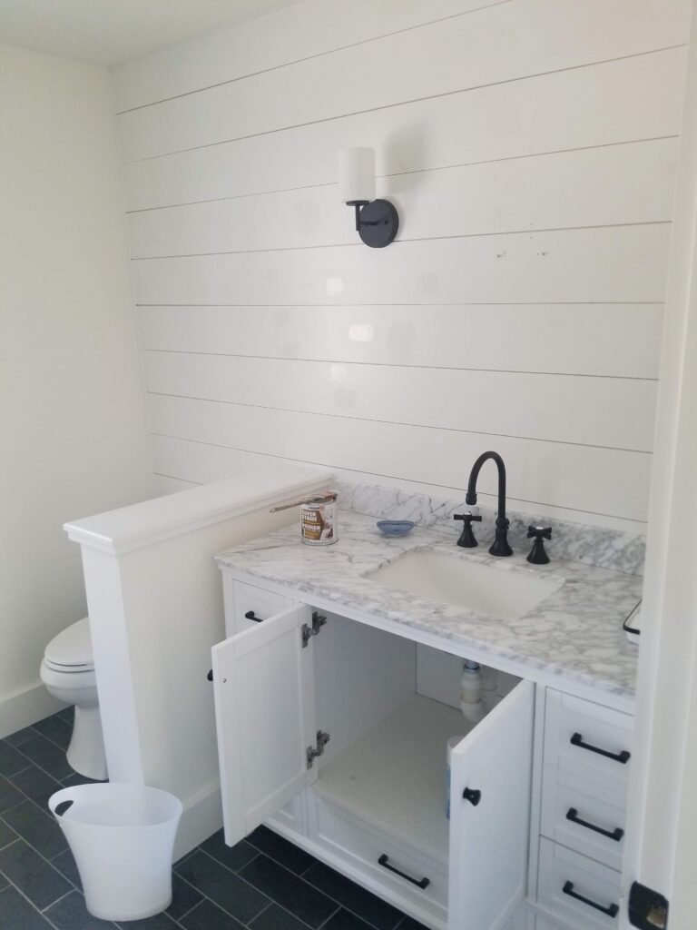 A modern bathroom with a half partition wall between the sink and toilet and marble top on the sink. The sink cabinets are white matching the tiles on the wall.