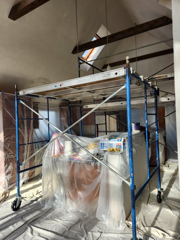 Inside a home, staging is set up over an area that has the homeowner's furniture condensed into a pile and covered with protective plastic film, and the floors are covered with drop cloths