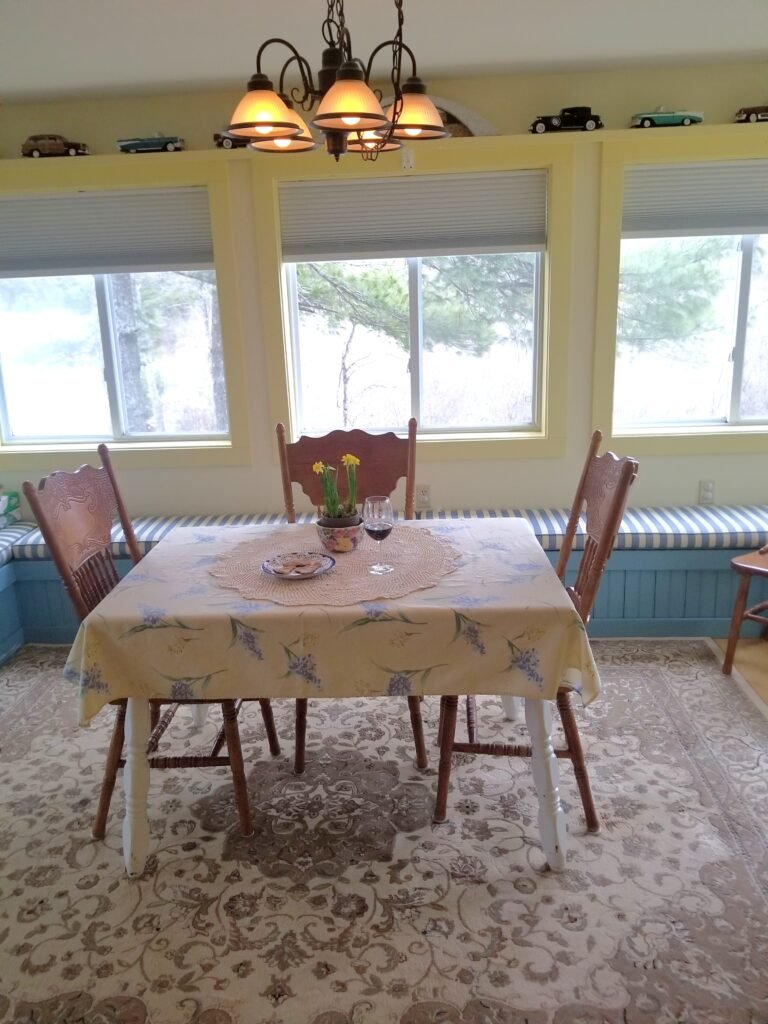A sunroom with three windows and a chandelier, a blue windowseat, and a small dining table. The walls have a yellow trim, and the tablecloth is yellow with blue accents to match the yellow and blue of the room.