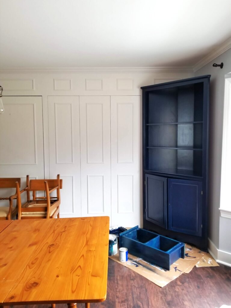An interior shot showing wood paneling with grooves that has been painted white with a corner cabinet freshly painted navy blue